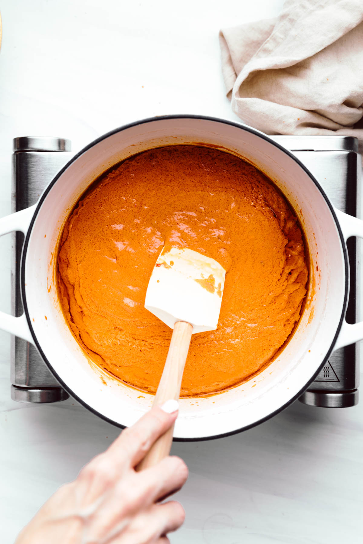 A large white pot with a thick orange colored sauce in it and a hand holding a spatula in the pot on a little stove with a napkin next to it.