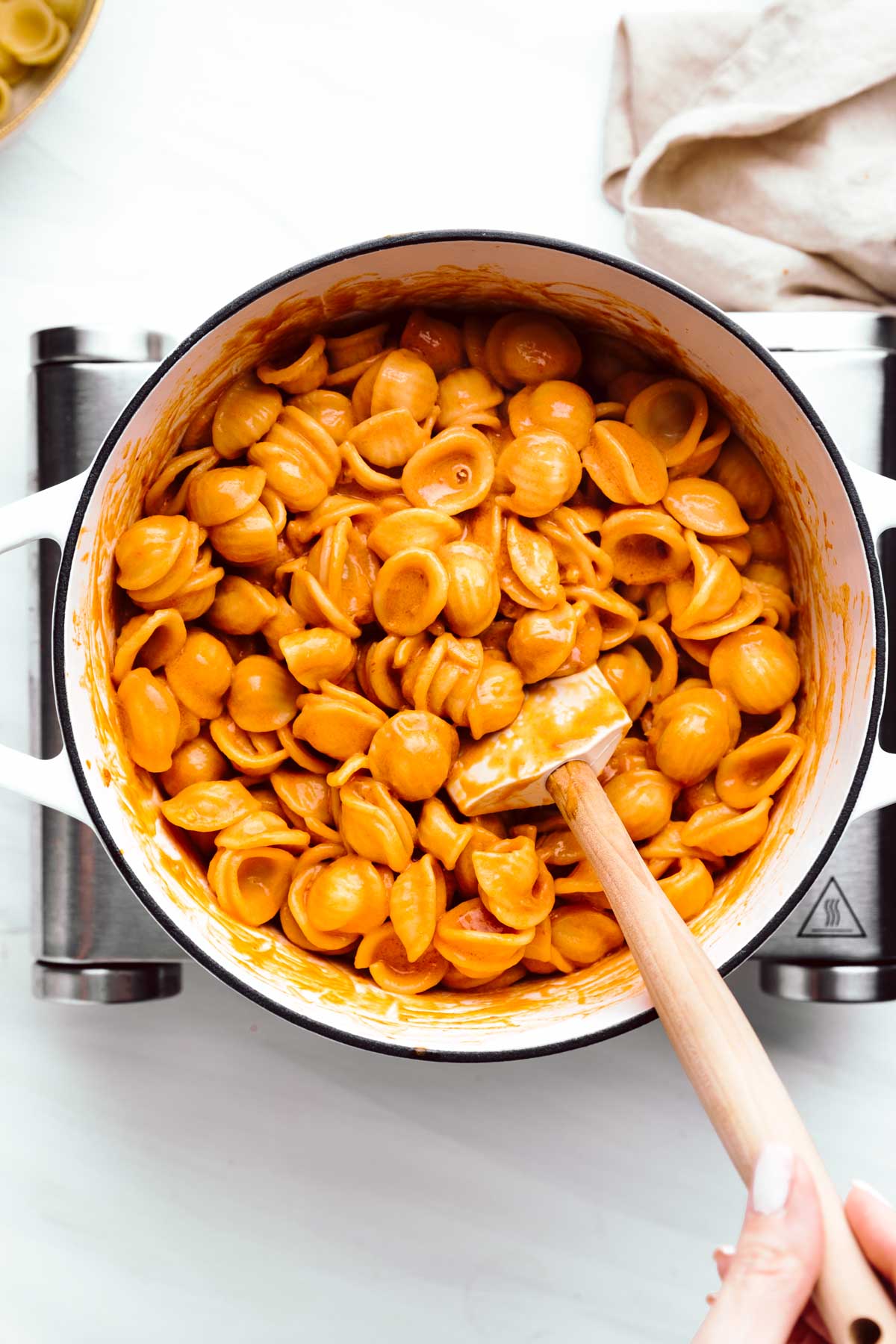 A large white pot with orange colored shell pasta and a spatula in the pasta held by a hand on a little stove with a napkin next to it.