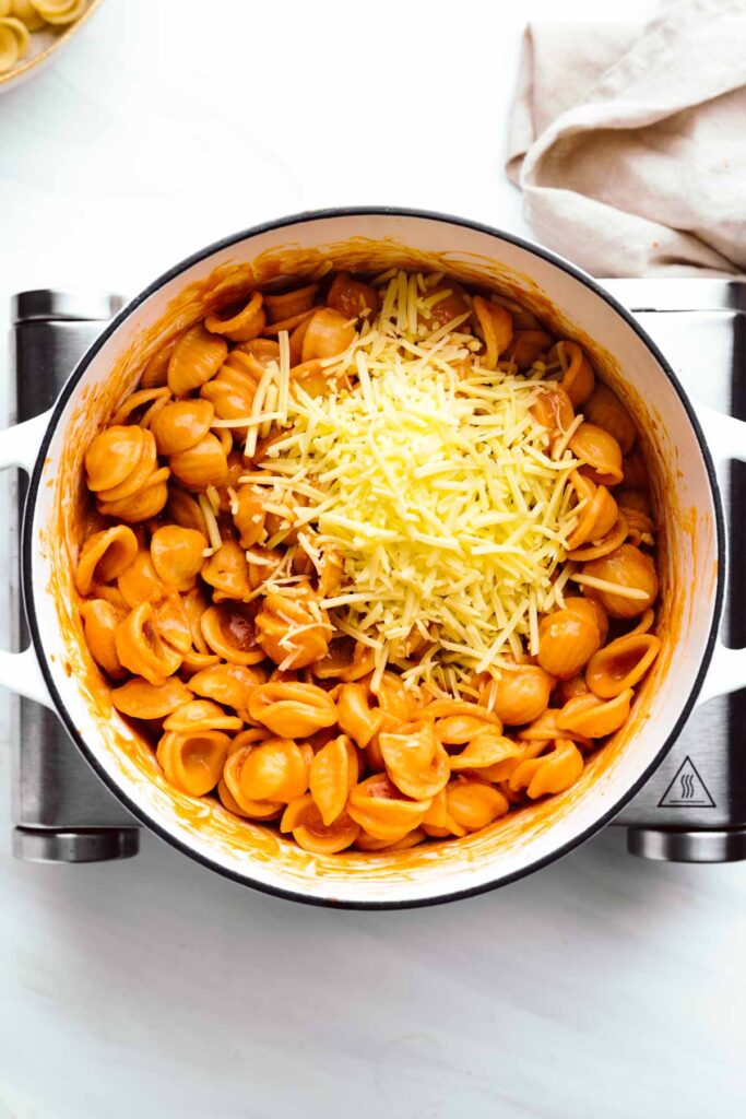 A large white pot with orange colored shell pasta in it topped with shredded Parmesan cheese on a little stove with a napkin next to it.