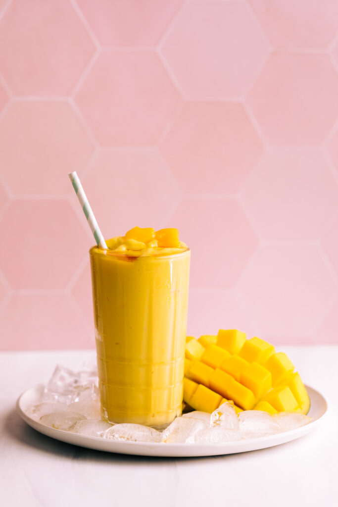 Mango lassi in a tall glass with a straw on a plate with ice cubes and fresh mango in front of a pink tile backdrop