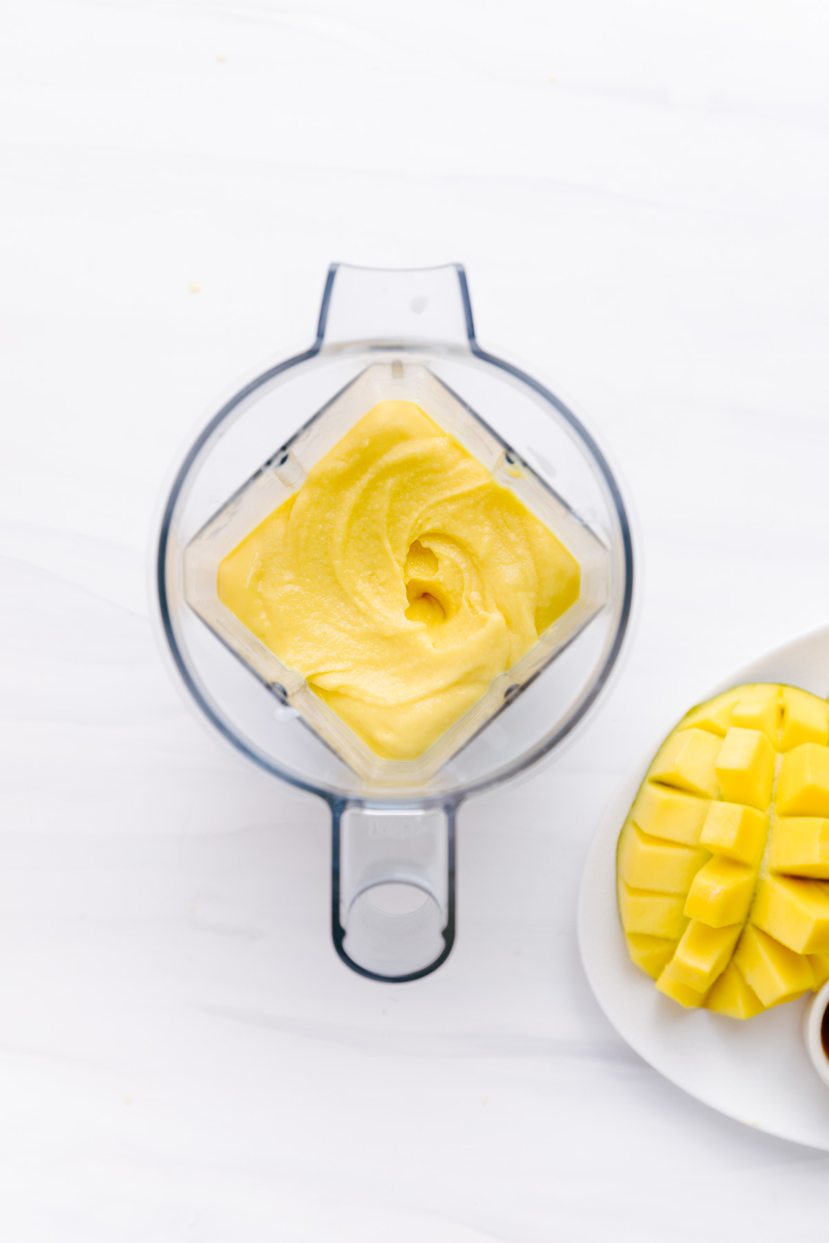 Blended ingredients for mango lassi in a blender on a white backdrop with fresh mango next to it