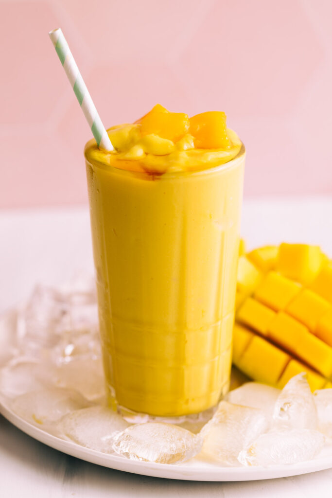 Mango lassi in a tall glass with a straw on a plate with ice cubes and fresh mango with a pink tile backdrop