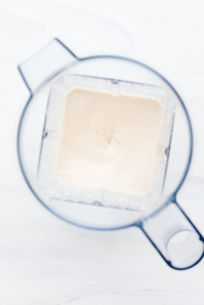 Top view of white bechamel sauce in a blender container on a white backdrop.