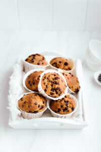 A white wooden tray with baked chocolate chip muffins placed on top of eachother in front of a white backdrop
