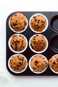 A black muffin tin pan with white cupcake liners and baked chocolate chip muffins in it
