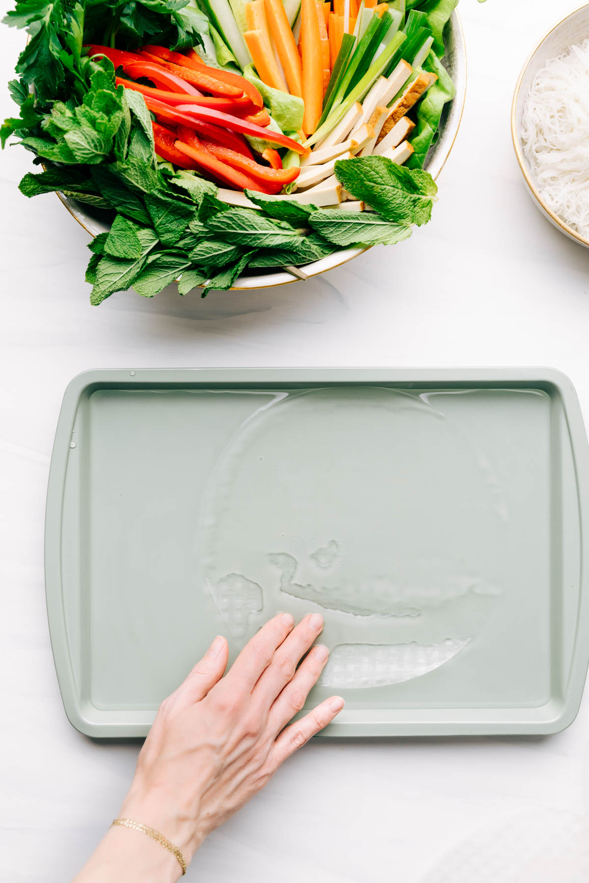 A light green baking tray filled with a bit of water and a hand emerging rice paper in it