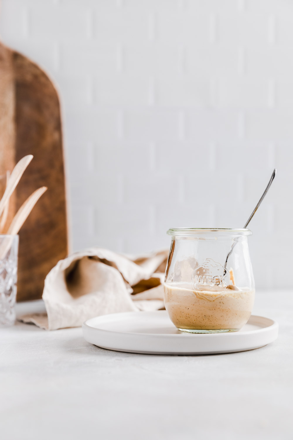 Spicy almond dressing in a small glass jar with a teaspoon on a light backdrop with a wooden cutting board, light brown napkin and wooden utensils in the background.