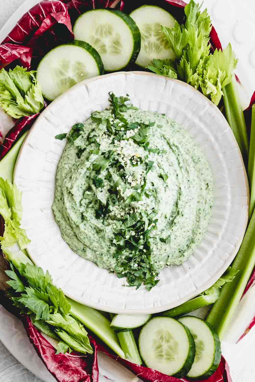 white bean dip in bowl with green herbs as topping on a backdrop of raw vegetables
