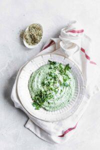white bean dip in a white bowl with green herbs as a topping on a light backdrop with a light napkin and a little bowl with hemp hearts