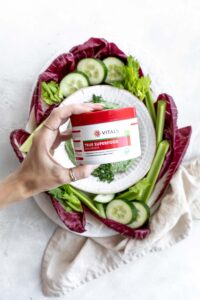 white bean dip in bowl with green herbs as a topping on a large white plate with raw vegetables on a white backdrop with a light brown napkin and a hand holding a jar of green superfood mix