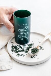 algro ritme spirulina with a hand holding the bottle on a white plate with spirulina sprinkles on a teaspoon