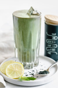 green smoothie in tall glass with ice cubes on white plate with a spoon with spirulina, lemon slices and mint leaves
