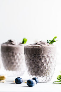 two glasses with blueberry smoothie in a small glass topped with mint leaves on a white marble cuttingboard with a white backdrop and a few blueberries