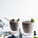 two glasses with blueberry smoothie in a small glass topped with mint leaves on a white marble cuttingboard with a white backdrop and a few blueberries in a small bowl and a napkin in the background