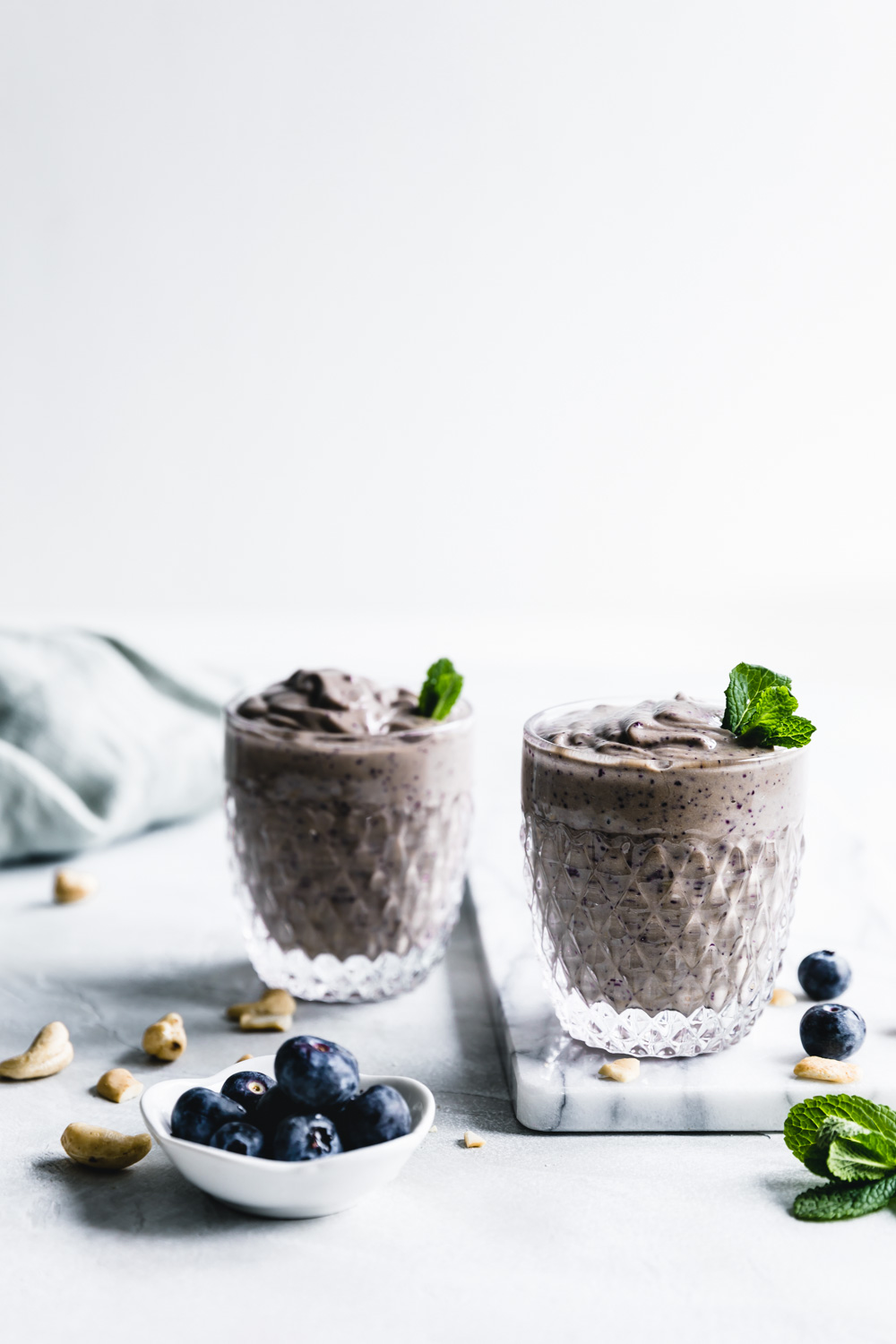 two glasses with blueberry smoothie in a small glass topped with mint leaves on a white marble cuttingboard with a white backdrop and a few blueberries in a small bowl and a napkin in the background