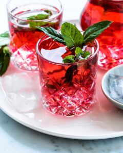 red cold hibiscus tea in glass with fresh mint leaves on white plate in front of a glass and a carafe with hibiscus tea