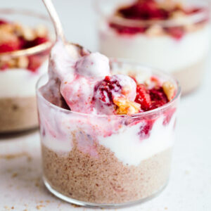 A small glass on a light backdrop with more puddings showing in the back layered with brown pudding, then white yogurt and then red raspberries with chopped walnuts and a spoon scooping up the yogurt with raspberries.