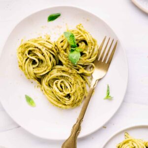 A light colored plate with three swirls of pasta al pesto on it and basil leaves as a garnish and a gold colored fork next to it with another pasta plate in the right corner.