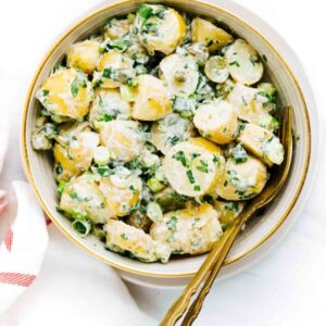 Potato salad with spring onions in a bowl with a gold fork and spoon on a white backdrop.
