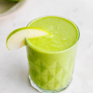 A green smoothie in a textured glass decorated with a slice of green apple on the rim of the glass on a round white marble backdrop cutting board with a green apple showing in the backdrop.