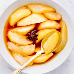 Stewed pears in an orange colored syrup in a white bowl with a star anise and a golden spoon on top of the pears.
