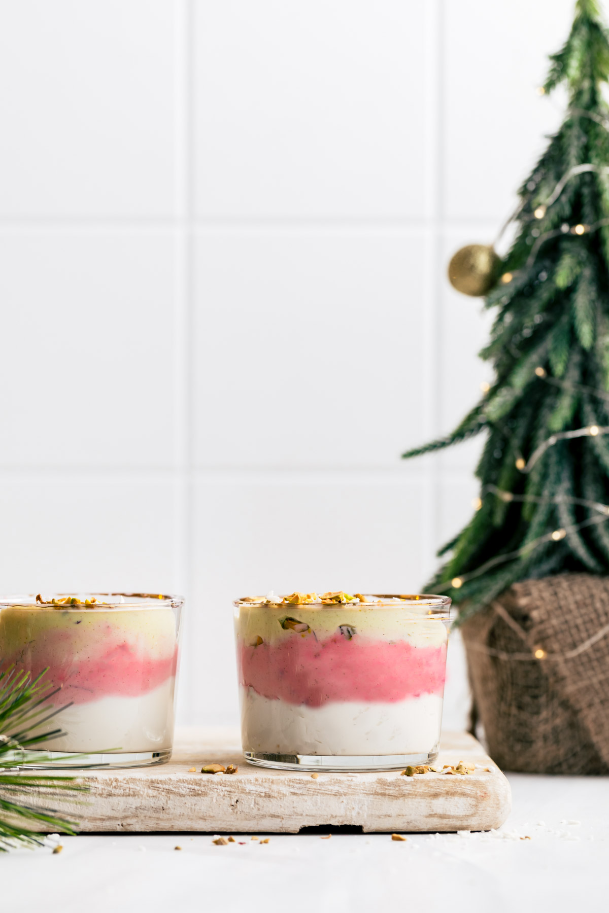 Two Christmas smoothies in a small glass topped with chopped pistachio nuts on a wooden cutting board in front of a small Christmas tree