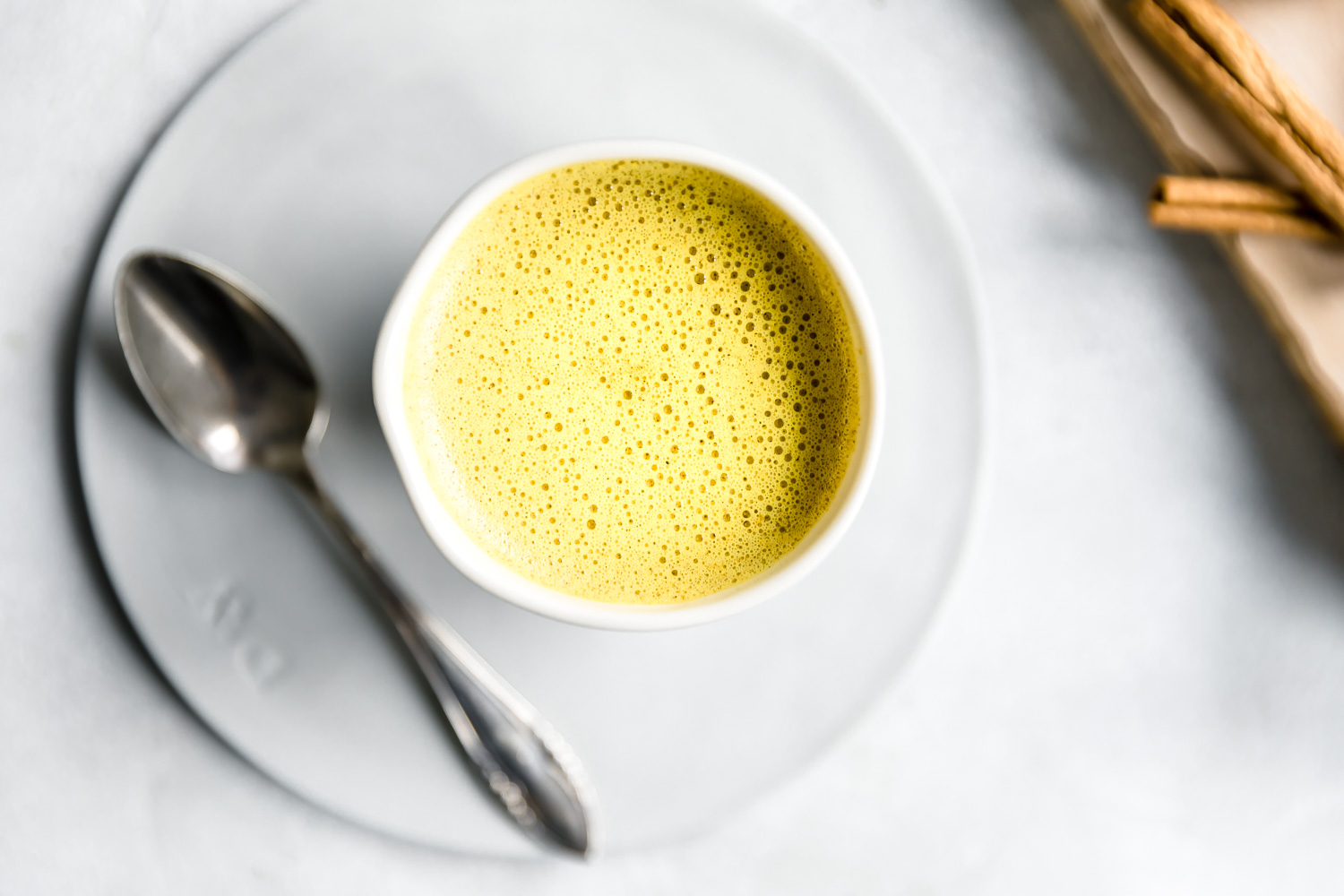 golden milk in white cup on a small white plate next to a silver teaspoon