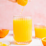 A glass with orange juice on a white table with a pink backdrop with oranges and more cups of orange juice next to it and a hand squeezing in more orange juice from an orange.