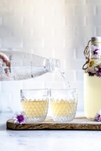 sodastream bottle pouring water in a small glass with tea next to another small glass with flowers and a tall bottle with tea and flowers on a wooden cutting board