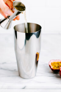 a silver colored cocktail shaker with orange passion fruit juice being poured into it