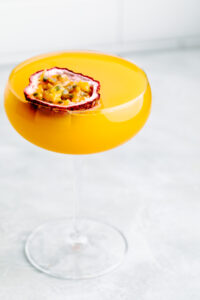 Orange colored drink Pornstar Martini Mocktail in a Martini glass with half a passion fruit floating on top on a light grey backdrop with white kitchen tiles in the background