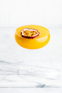Orange colored drink Pornstar Martini Mocktail in a Martini glass with half a passion fruit floating on top on a white marble cutting board with white kitchen tiles in the background
