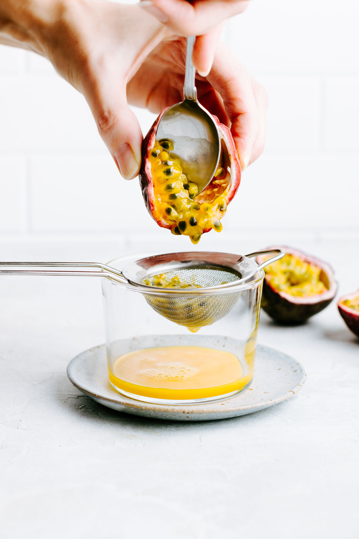a small glass on a blue plate with a small sieve hanging in the glass and a hand scooping out the seeds and pulp form the passion fruit with a teaspoon in the sieve