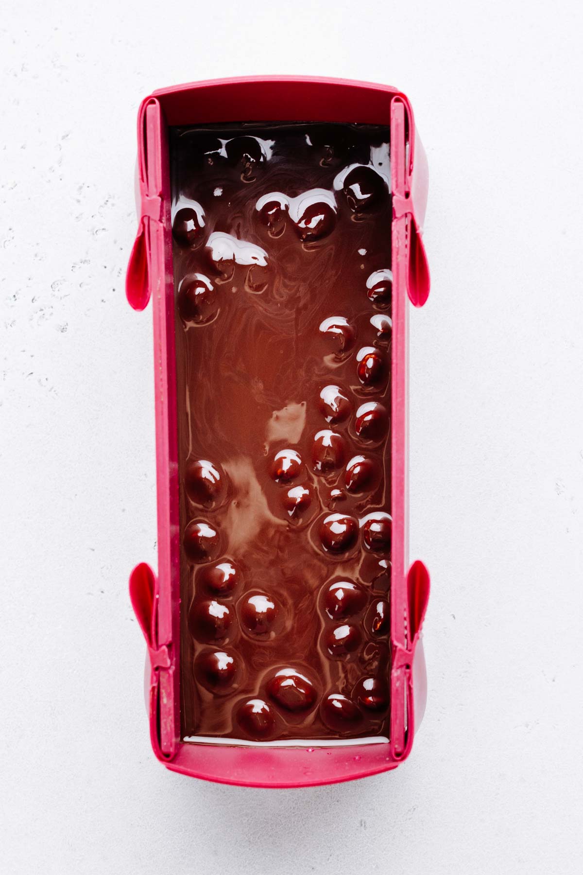 a burgundy colored silicone loaf pan with melted chocolate and hazelnuts in it on a white marble backdrop.