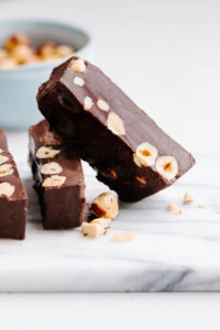 three brown colored chocolate torrone slices with hazelnuts chunks showing on the inside of the slices and a few hazelnuts next to it on a white marble cutting board.