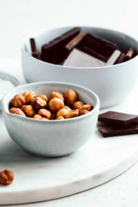 a small blue bowl with hazelnuts on a marble cutting board and a large blue bowl with chocolate chunks in the back.