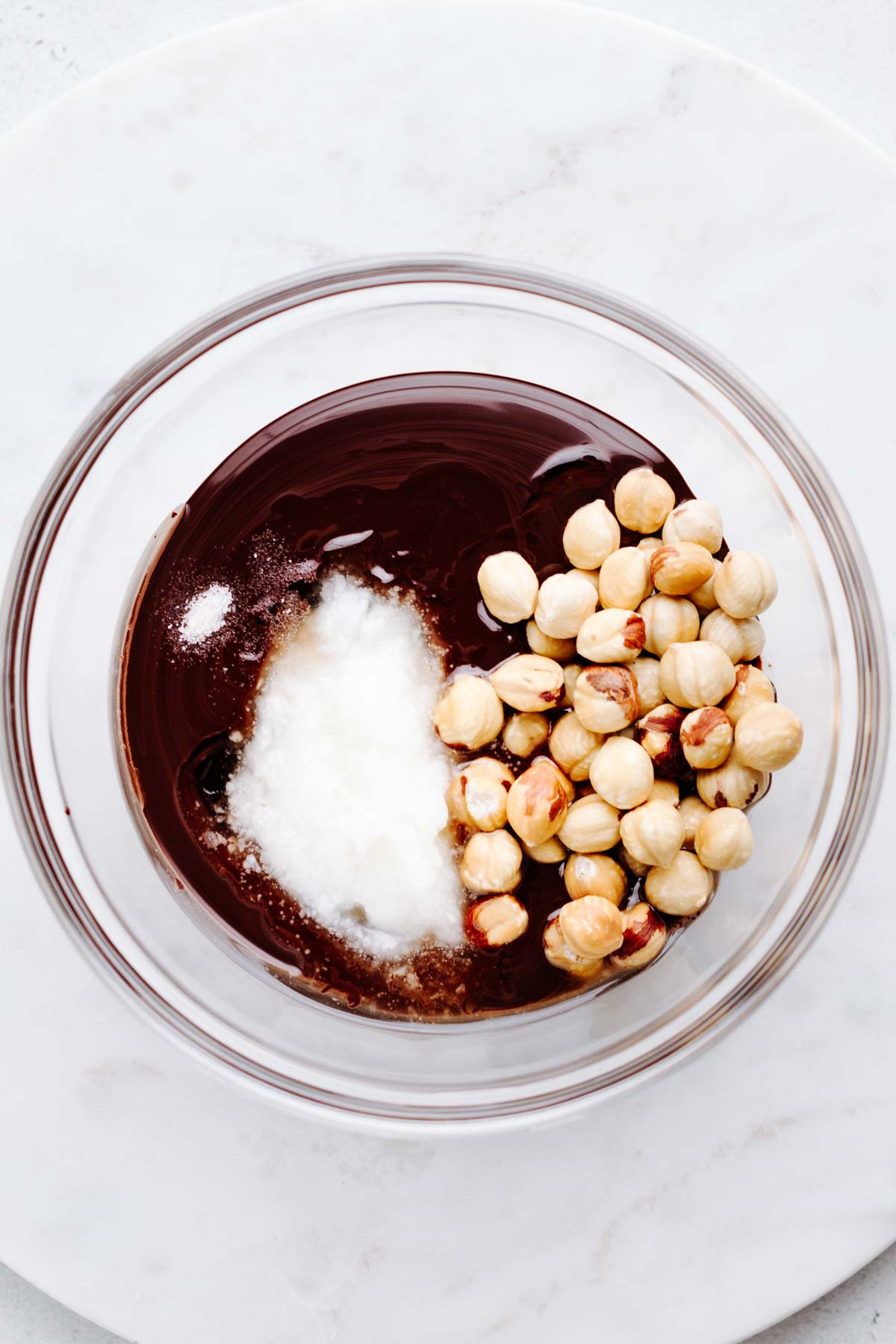 a transparent bowl on a white marble backdrop with melted chocolate, coconut oil, salt, maple syrup and hazelnuts in it.