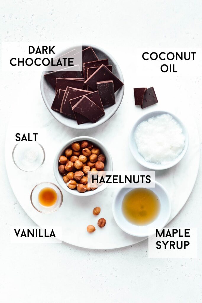 the ingredients for chocolate torrone on a round marble cutting board in small bowls and with the name of the ingredients next to it.